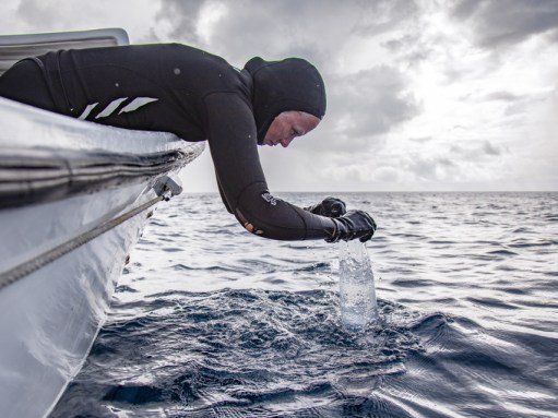 a diver leans out over the edge of a boat and scoops water into a bag