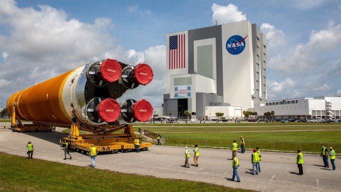 A huge rocket booster on its side, resting on two wheeled platforms, in front of a large blocky building. Workers in yellow vests walk nearby.